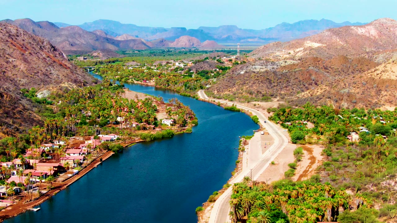 Wide aerial river valley with road, palms, buildings, dry hills, and mountains.
