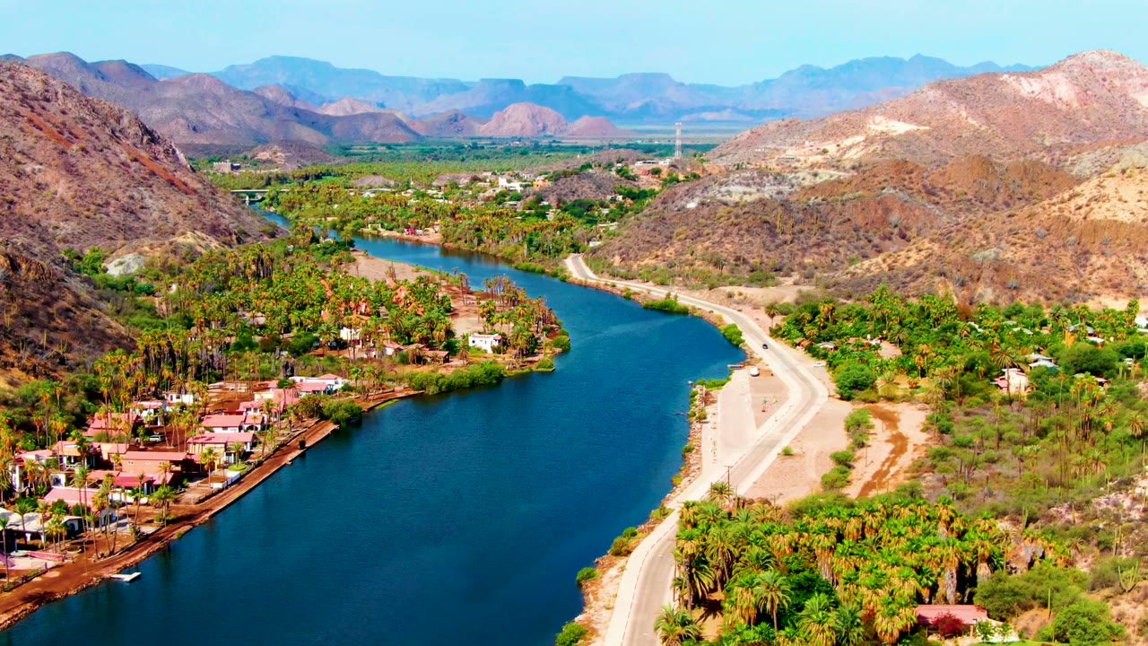 Mid-clip aerial view over Mulege, Baja California Sur with the river bend shifted right, a pale roadside bank, green palms, clustered buildings, dry hills, distant mountains, and no visible people.