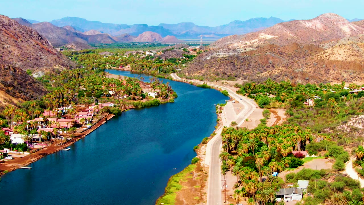 Opening aerial frame over Mulege, Baja California Sur, showing a blue river, roadside palms, dry hills, scattered buildings, and no visible people.
