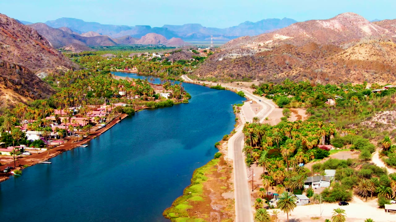 Mulege aerial frame with a broad blue river bend, palms and homes along the water, the road tracing the opposite bank, and no visible people.
