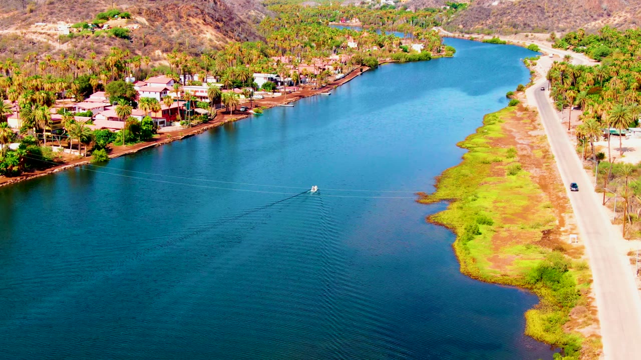 Aerial view in Mulege, Baja California Sur, showing blue canal water, a pale road on the right, waterfront homes and palms on the left, dry hills beyond, and no visible people.