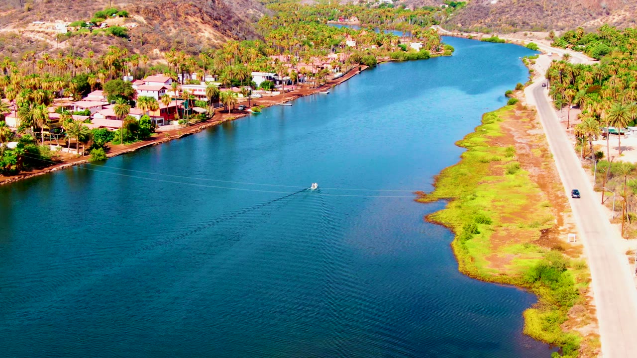 Small boat crossing a canal beside a road and residential shoreline.