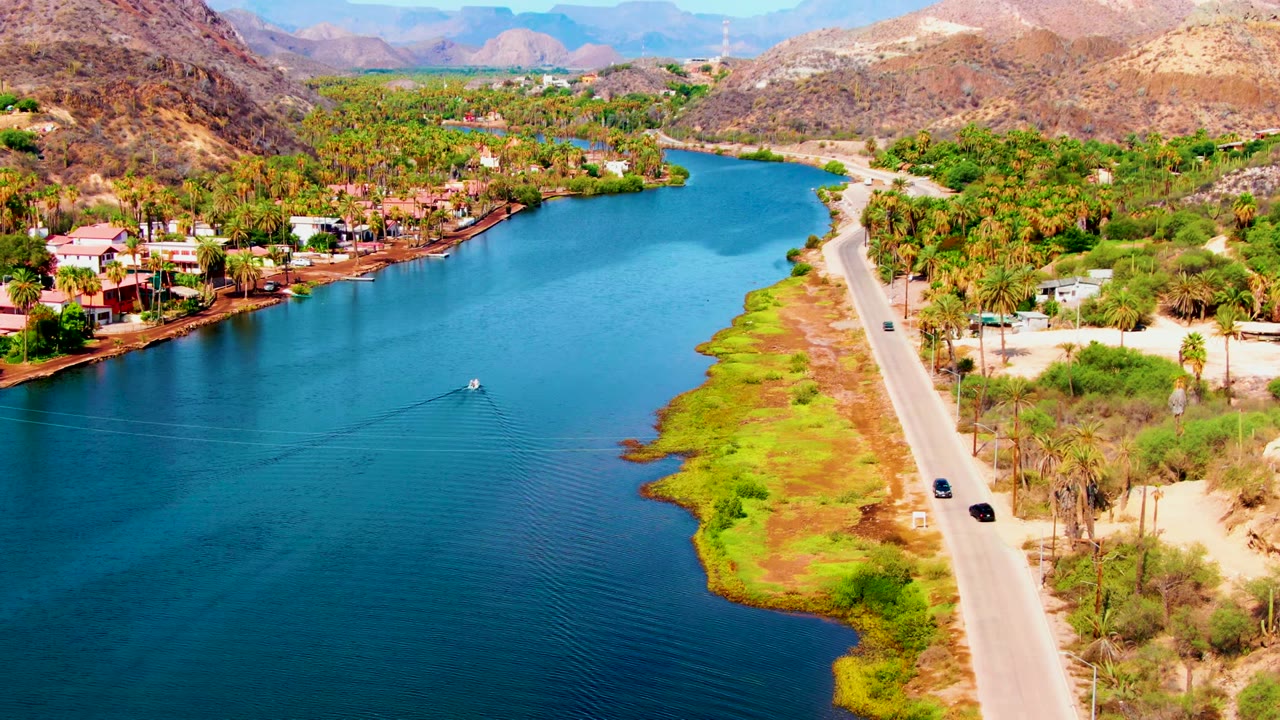 Aerial view over Mulege, Baja California Sur, showing a blue canal beside a pale road, palm trees, shoreline houses, dry hills, distant mountains, and no visible people.
