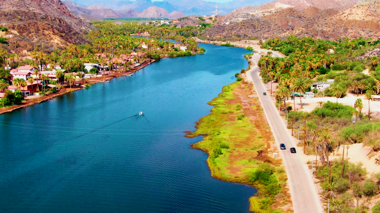Drone view of a canal bend with a road, cars, palms, houses, and mountains.