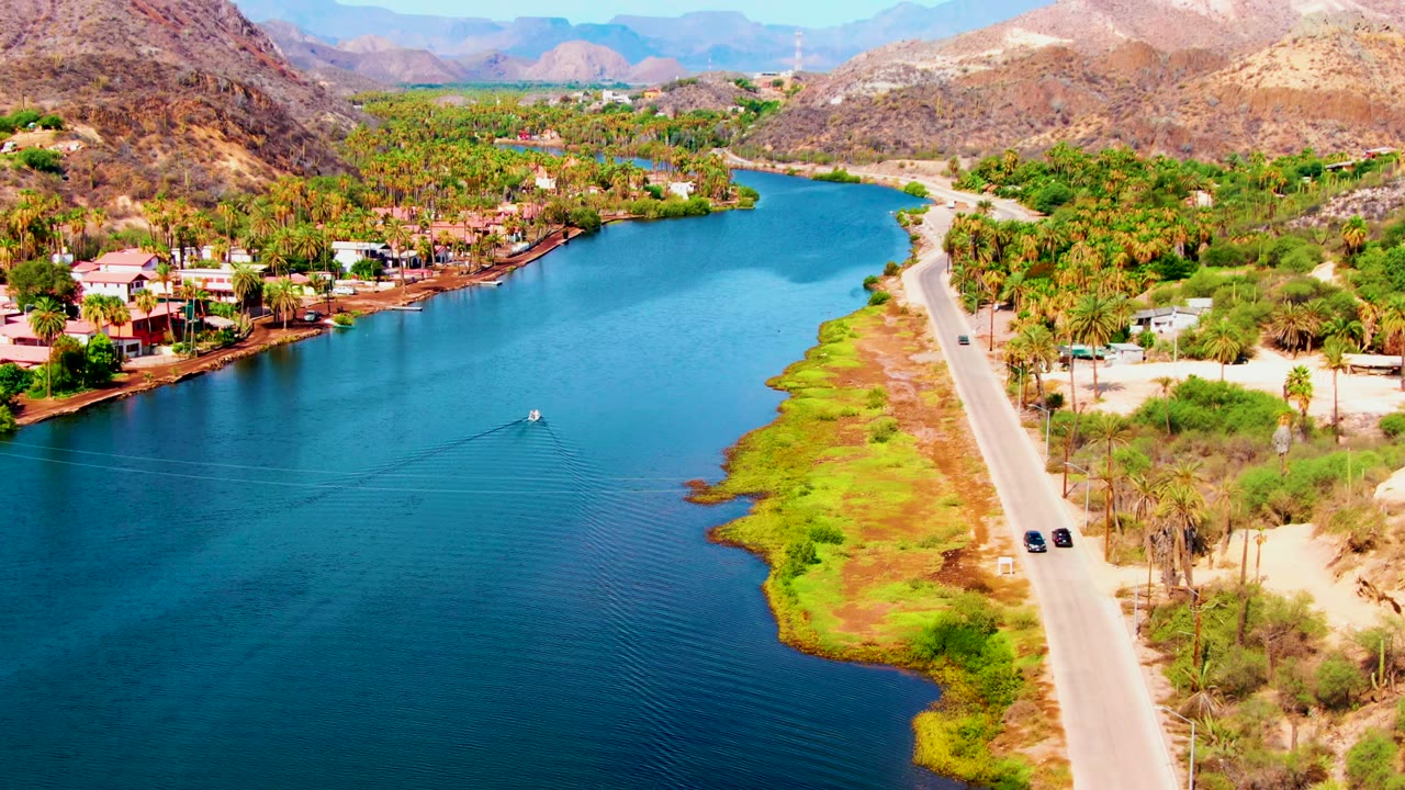 Wide aerial over Mulege, Baja California Sur, showing two cars on the road beside the canal, a small boat wake on the blue water, palm clusters, shoreline buildings, dry hills, and no visible people.