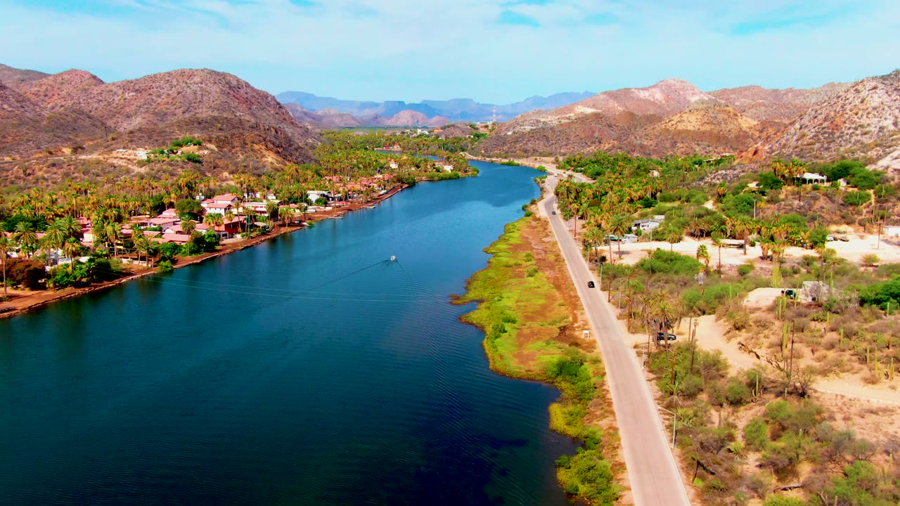 Aerial view in Mulege, Baja California Sur, with a blue river beside a pale road, houses among palms and dry brush, rocky hills beyond, and no visible people.