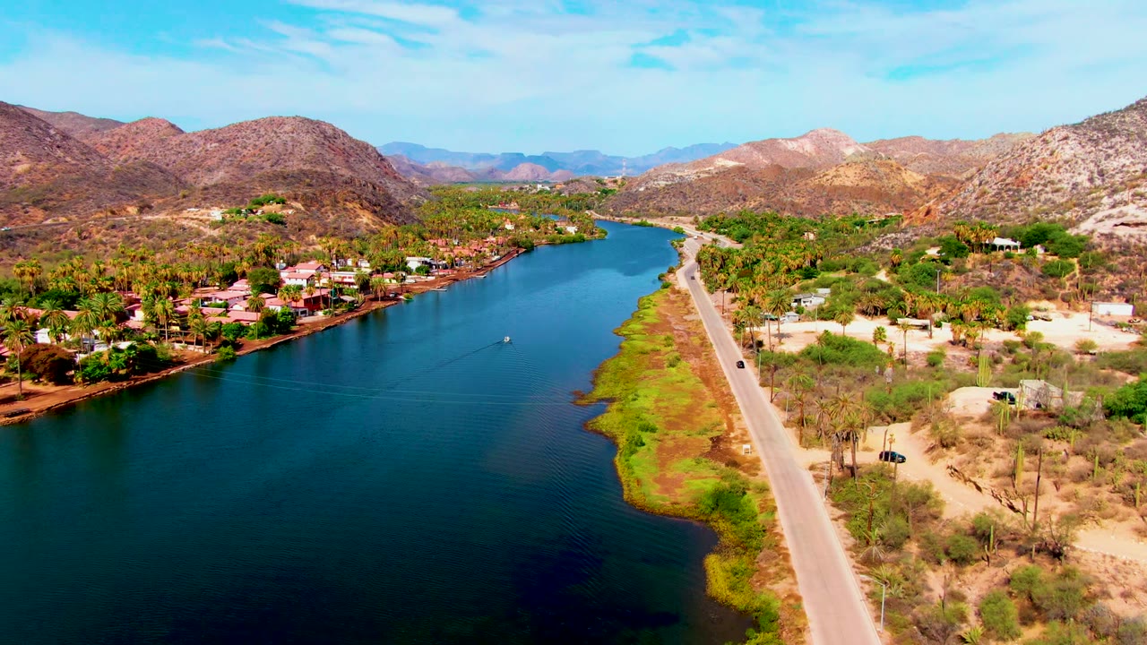 Aerial view in Mulege, Baja California Sur, with a small vehicle on the road beside the blue river, homes and palms near the water, brown hills behind, and no visible people.