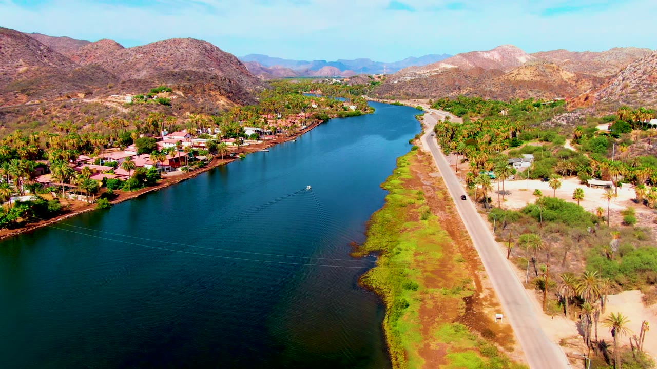 Aerial view over Mulege, Baja California Sur, with a blue river, roadside vegetation, houses along the bank, desert mountains in the background, and no visible people.