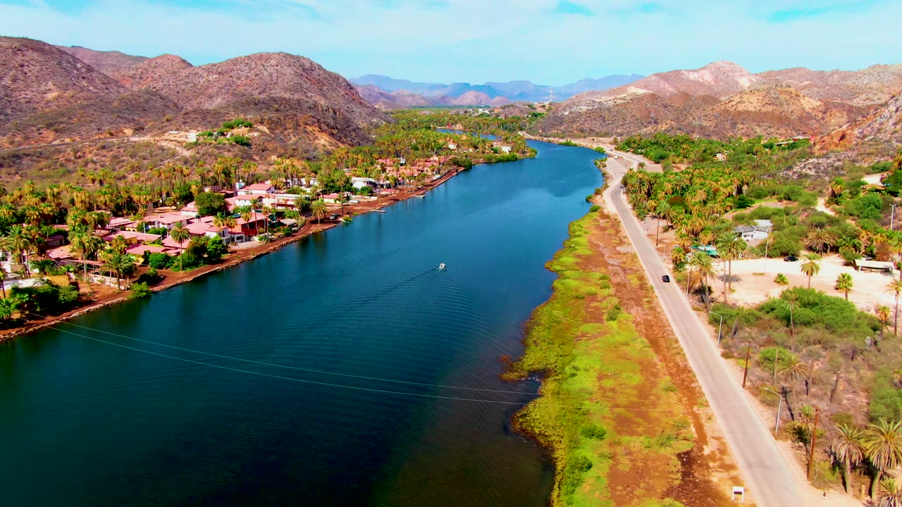 Wide aerial frame of the river curving past homes, palms, and a roadside.