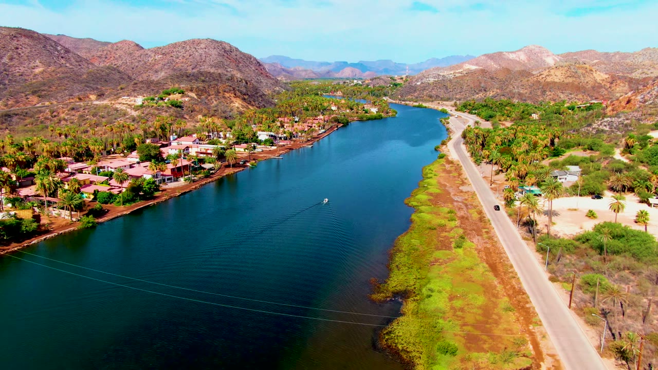 Drone-height view in Mulege with the river filling the left side, a pale road on the right, scattered homes and palms near the bank, mountain ridges behind, and no visible people.