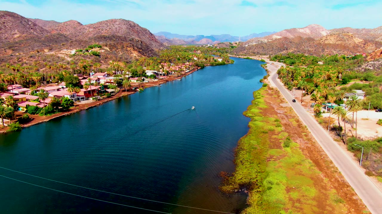 Aerial view in Mulege, Baja California Sur, showing a blue-green waterway beside a shoreline road, residential homes, palms, dry hills, and no visible people.