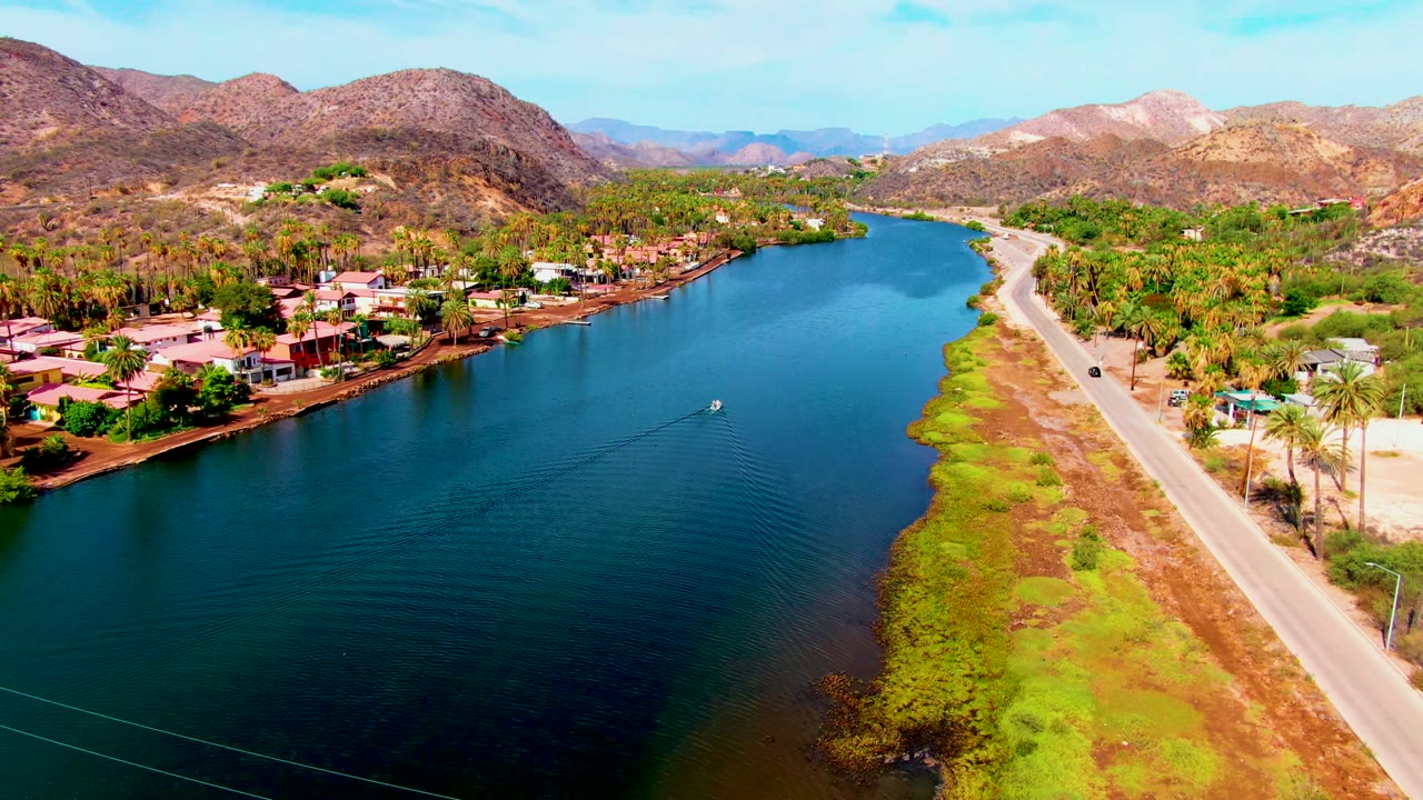 Aerial Mulege frame with the small boat farther across the water, ripples spreading behind it, a car on the road, homes on the bank, and no visible people.