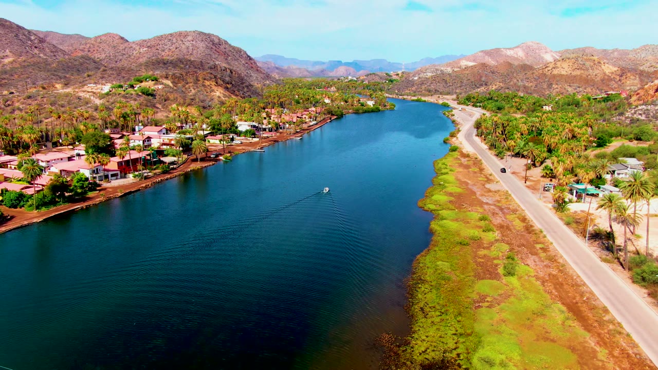 Aerial view over Mulege with the boat near midwater, the road running along the right shore, clustered homes and palms on the left, mountains behind, and no visible people.