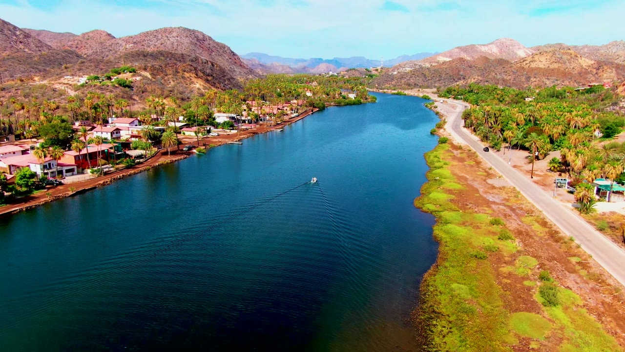 Aerial frame from Mulege, Baja California Sur, looking along blue water with shoreline homes on the left, a road on the right, dry hills in the background, and no visible people.