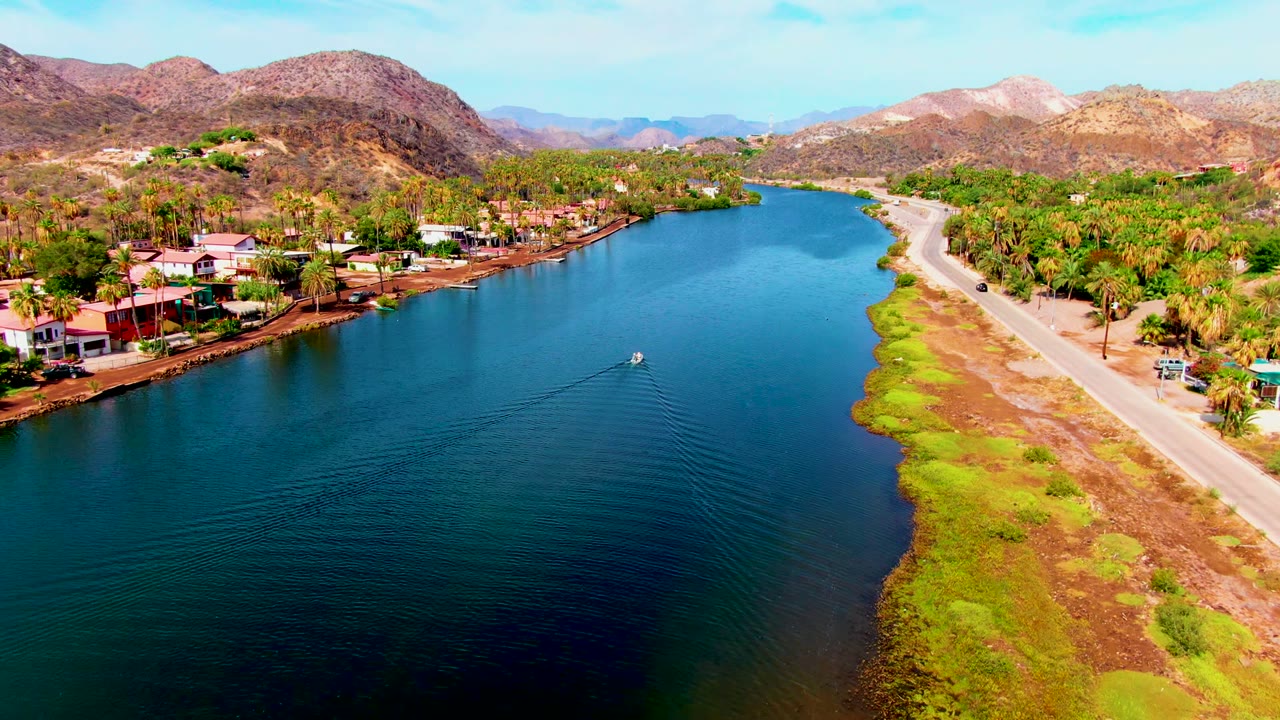 Mulege aerial frame showing the boat farther along the water with widening ripples, the pale road following the right shoreline, clustered homes on the left, and no visible people.