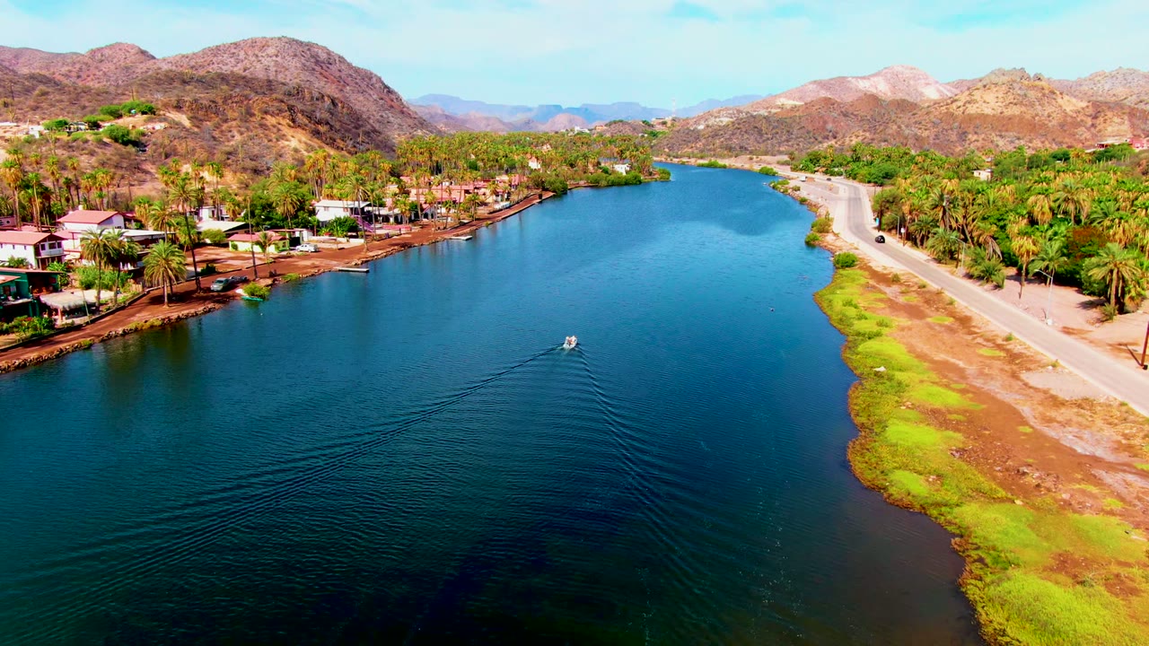 Aerial view in Mulege, Baja California Sur, with a small white boat moving across blue canal water, a road on the right, palm-lined houses on the left, dry mountains beyond, and no visible people.