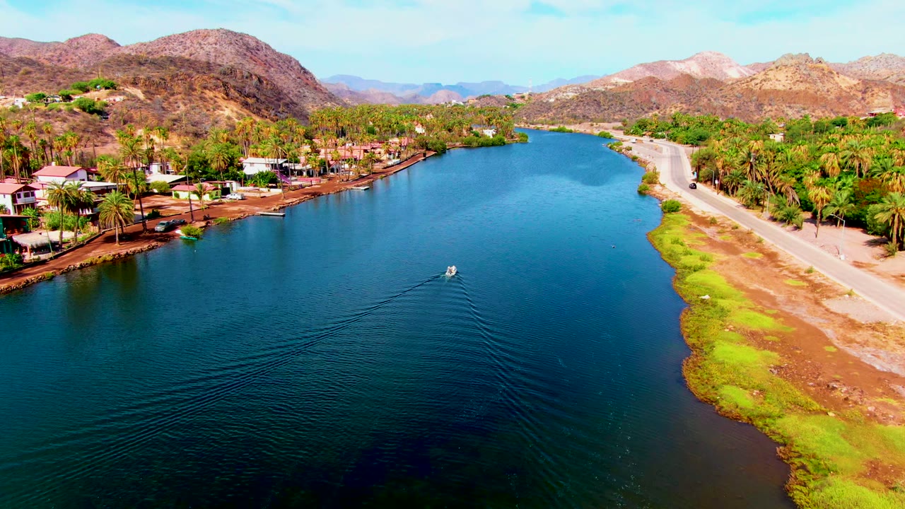 Boat wake spreads across blue water near a road, palms, houses, and mountains.