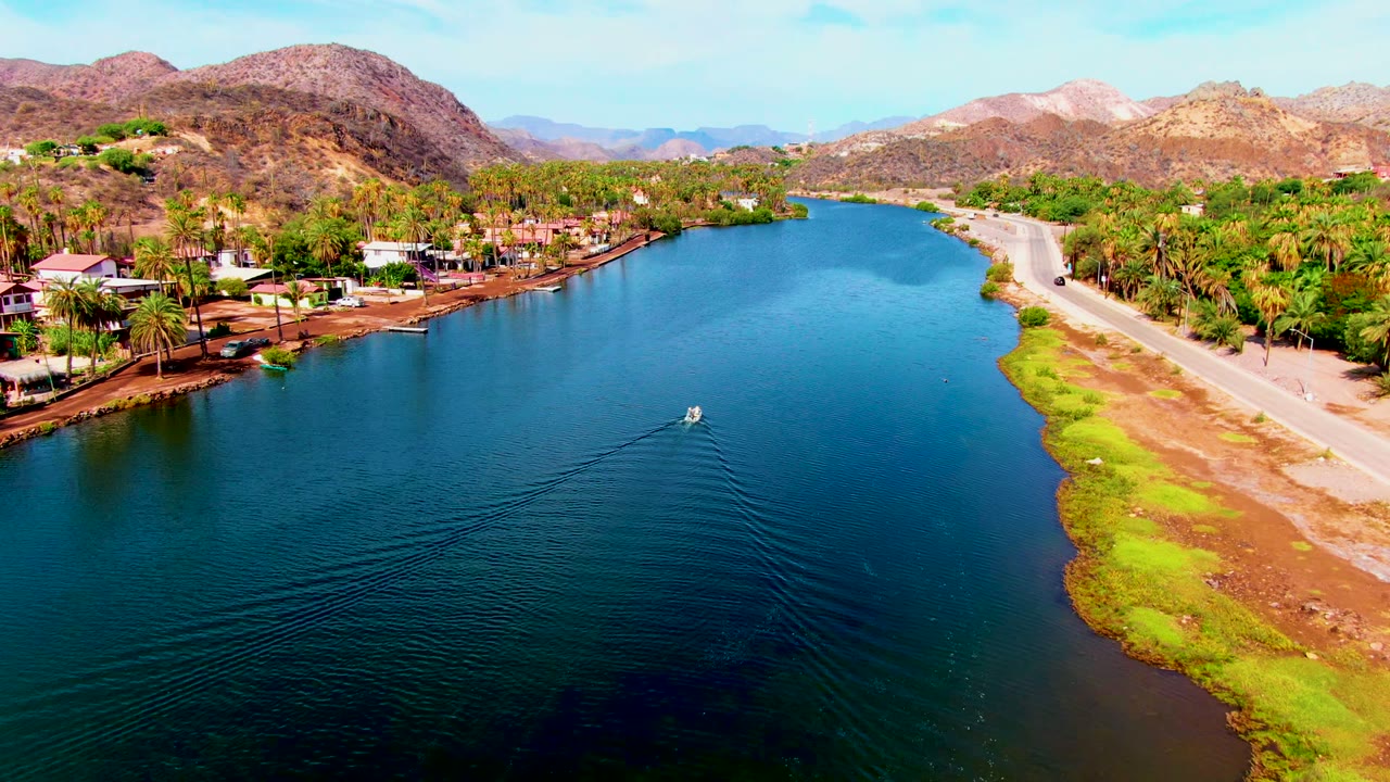 Midway through the aerial pass in Mulege, the boat is visible near the center-right of the canal, its wake trailing over dark blue water, with homes, palms, a pale road, dry hills, and no visible people.