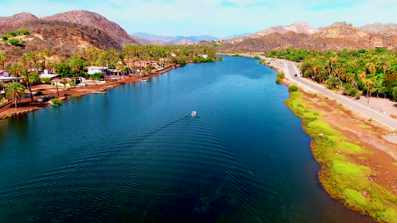 Aerial frame from Mulege, Baja California Sur showing a small boat beginning its path across blue canal water beside a road, palms, low buildings, dry hills, and no visible people.