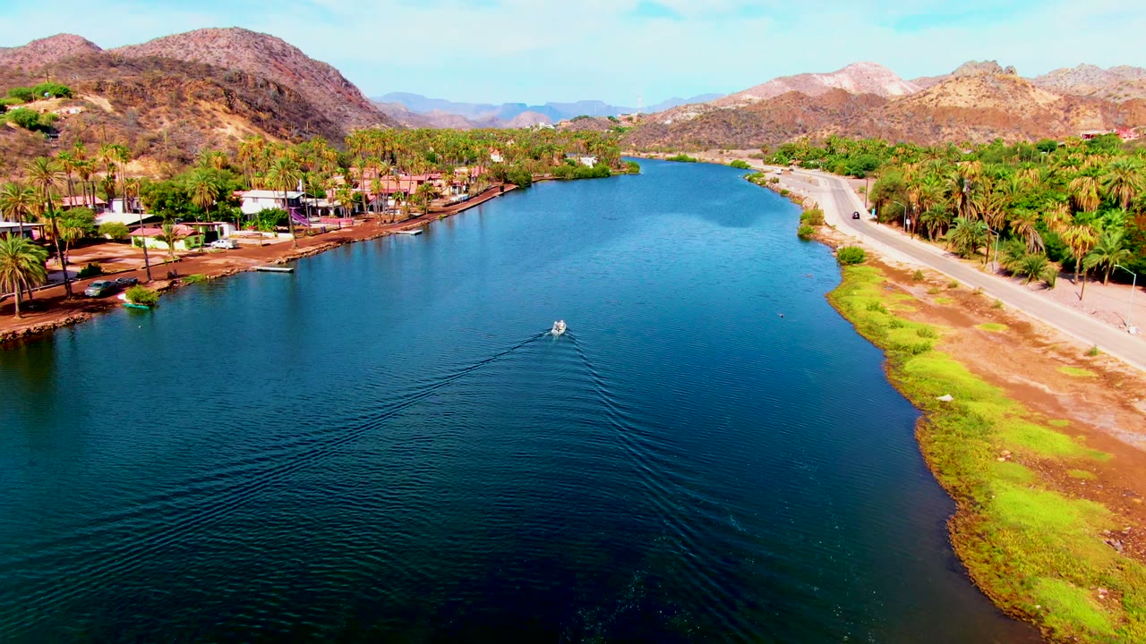 Small boat leaves a wake in canal water near a road and green shoreline.