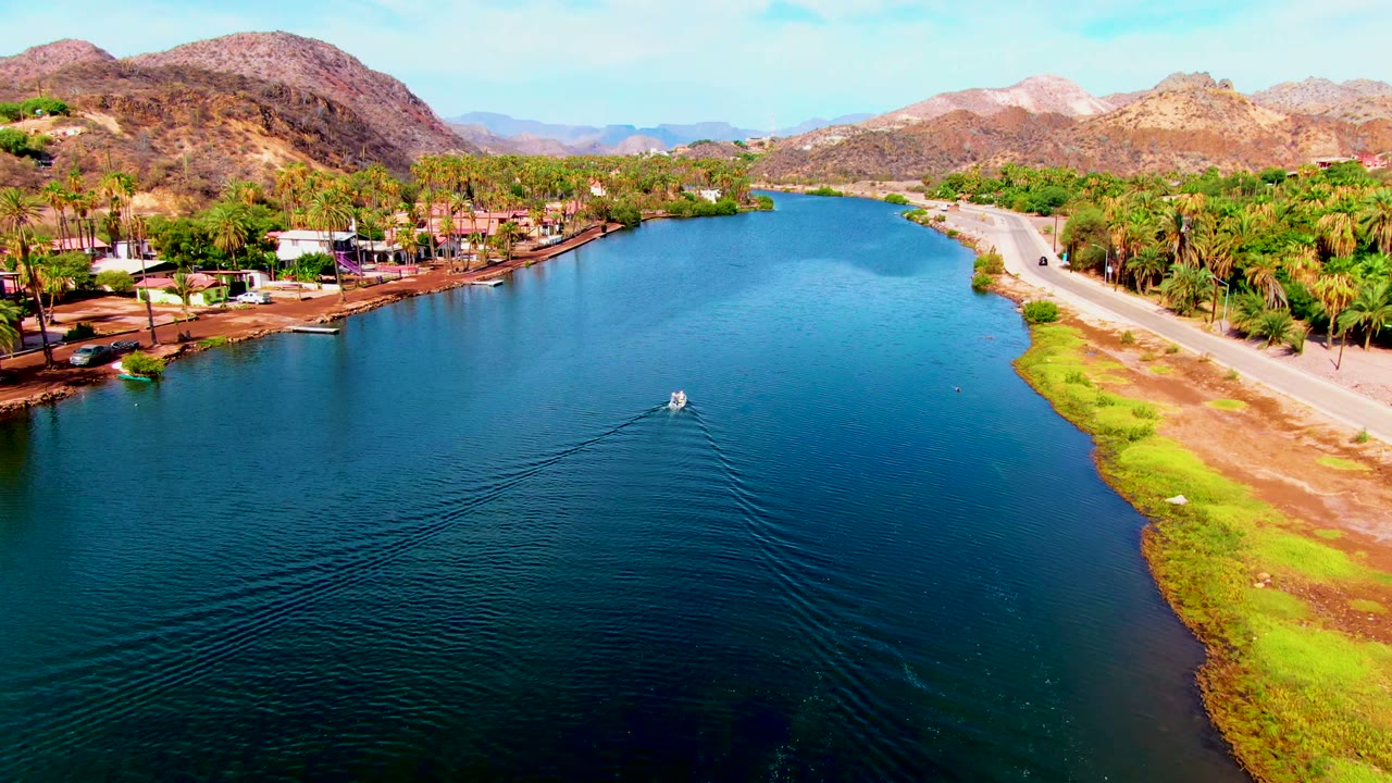 The boat continues along the Mulege canal as the wake widens across darker foreground water, with palms, roadside land, low buildings, mountains, and no visible people.