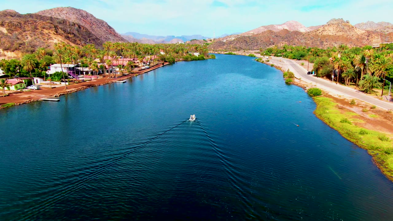 Early aerial view in Mulege, Baja California Sur shows a small boat beginning across blue canal water, with palm trees, low buildings, a roadside bank, brown mountains, and no visible people.