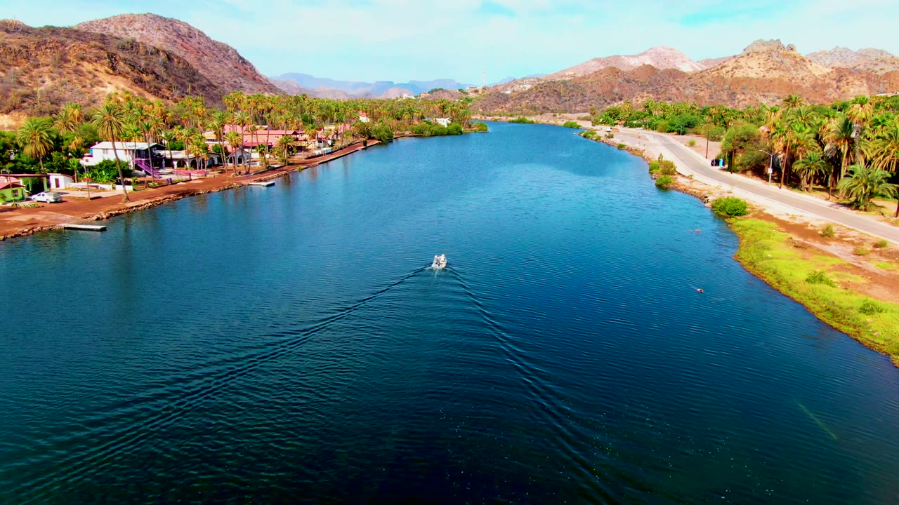 Mid-clip aerial view in Mulege, Baja California Sur keeps the small boat near the center of the canal, with ripple lines behind it, a road on the right bank, palms and buildings on the left, and no visible people.