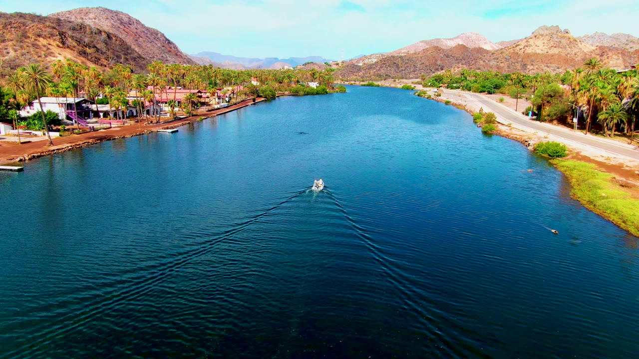 Aerial view in Mulege, Baja California Sur of a small white boat moving along a blue canal, with palm trees and low buildings on the left bank, a road on the right bank, mountains beyond, and no visible people.