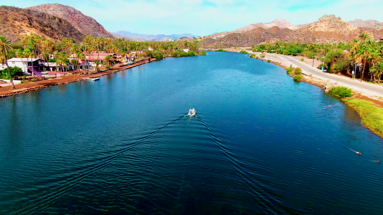 Elevated view in Mulege, Baja California Sur with the boat continuing up the canal, a broad wedge of ripples behind it, palm trees and buildings along one shore, a pale road along the other, and no visible people.