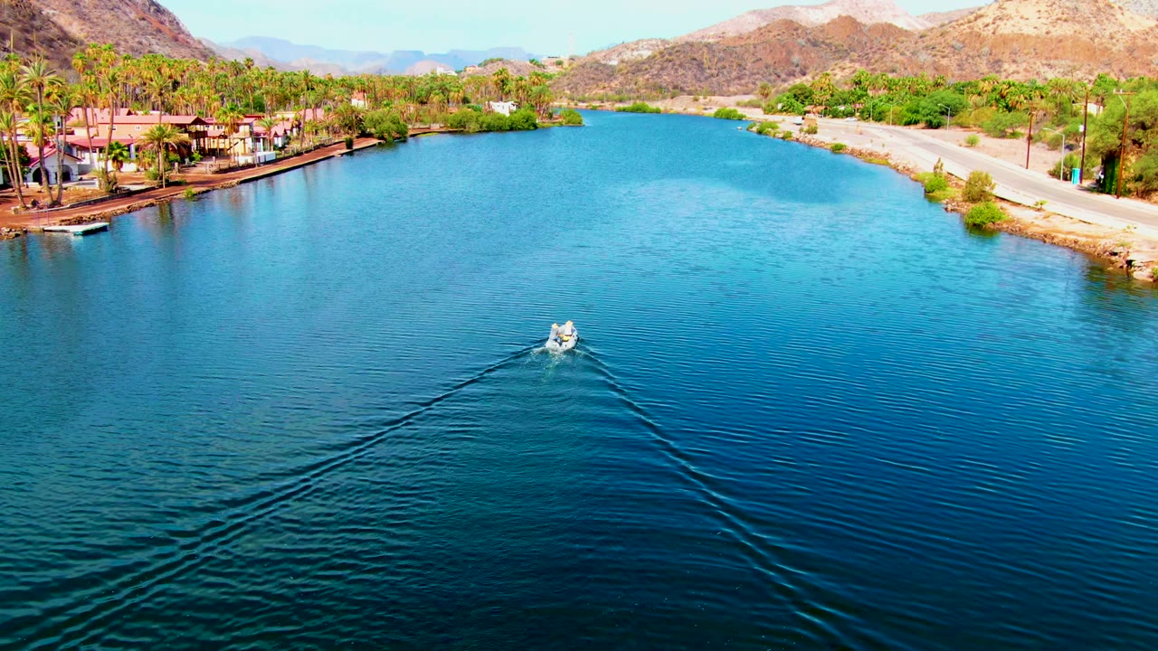 Aerial view in Mulege, Baja California Sur of a small white boat moving away across blue canal water, with palm trees, waterfront buildings, a road, tan hills, and no visible people.