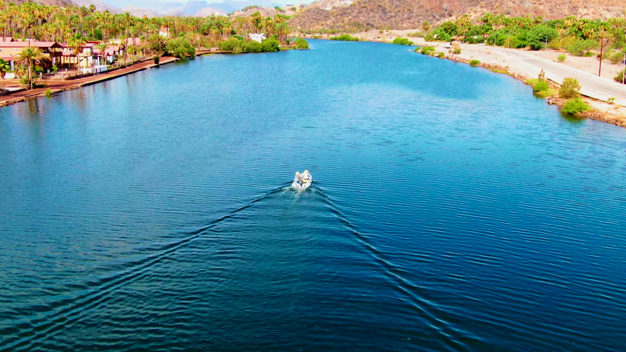 High aerial view over the Mulege canal as a small white boat creates a long V-shaped wake on blue water, with shoreline palms, buildings, road, greenery, hills, and no visible people.