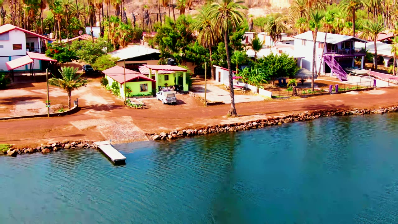 Aerial frame over Mulege, Baja California Sur showing blue rippled water, a small dock, red-roofed waterfront homes, palms, parked vehicles, and no visible people.
