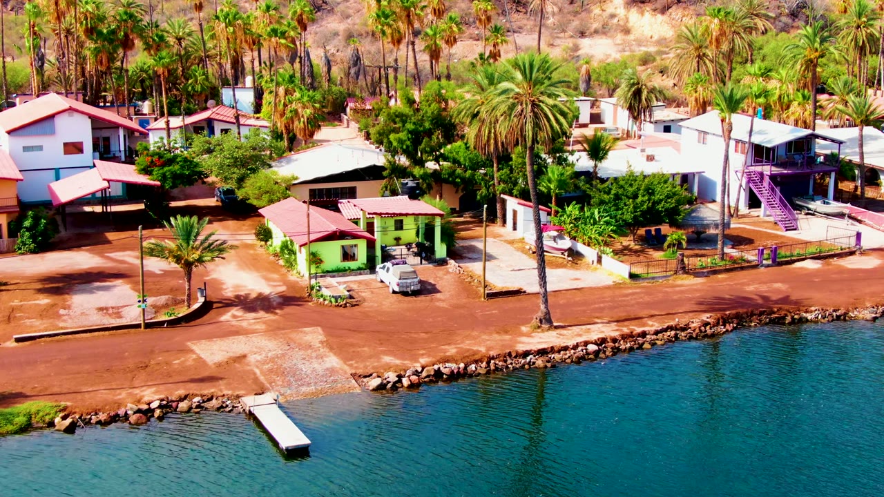 Aerial frame over Mulege, Baja California Sur showing the green waterfront house and parked vehicle more prominently above the rocky shore, with rippled blue water and no visible people.