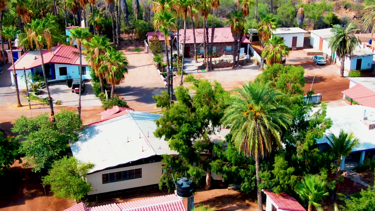 Opening overhead frame of a Mulege, Baja California Sur residential block with tall palms, red and white roofs, dirt lanes, a blue car near a white building, and no visible people.