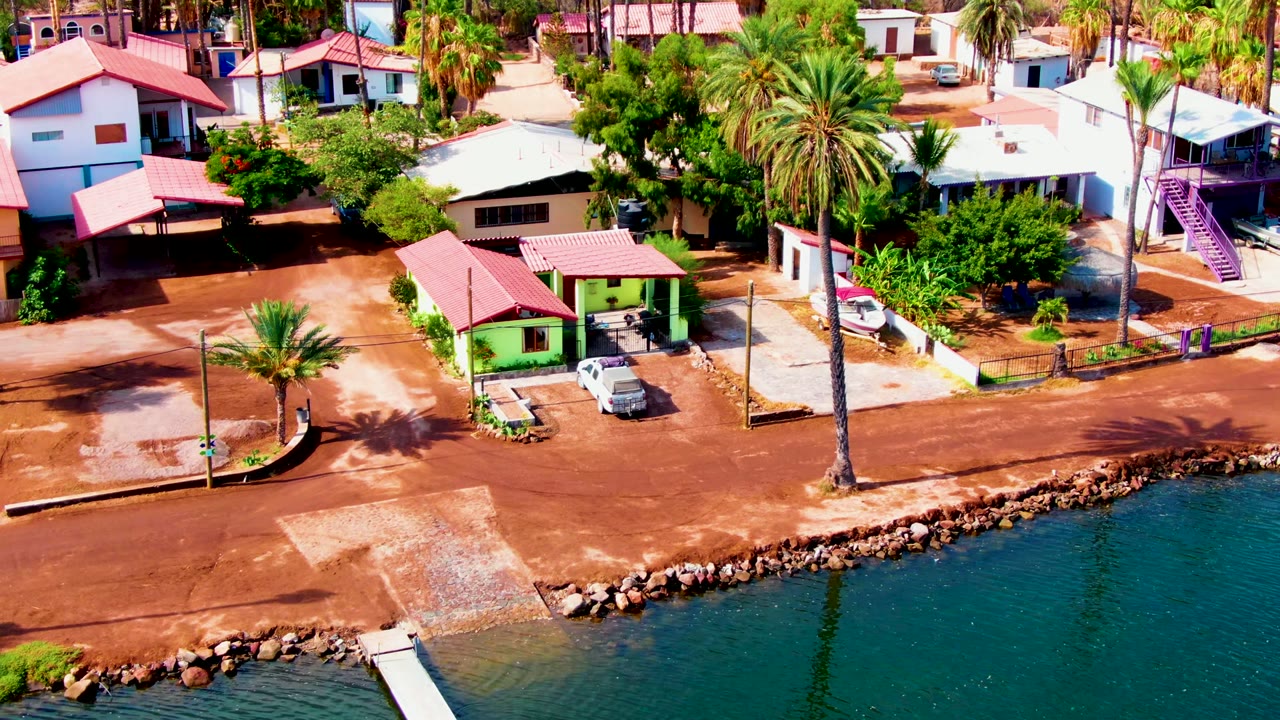 Aerial frame in Mulege, Baja California Sur showing blue-green water and a small dock at the lower edge, a reddish dirt street, red-roofed homes, palms, parked cars, and no visible people.