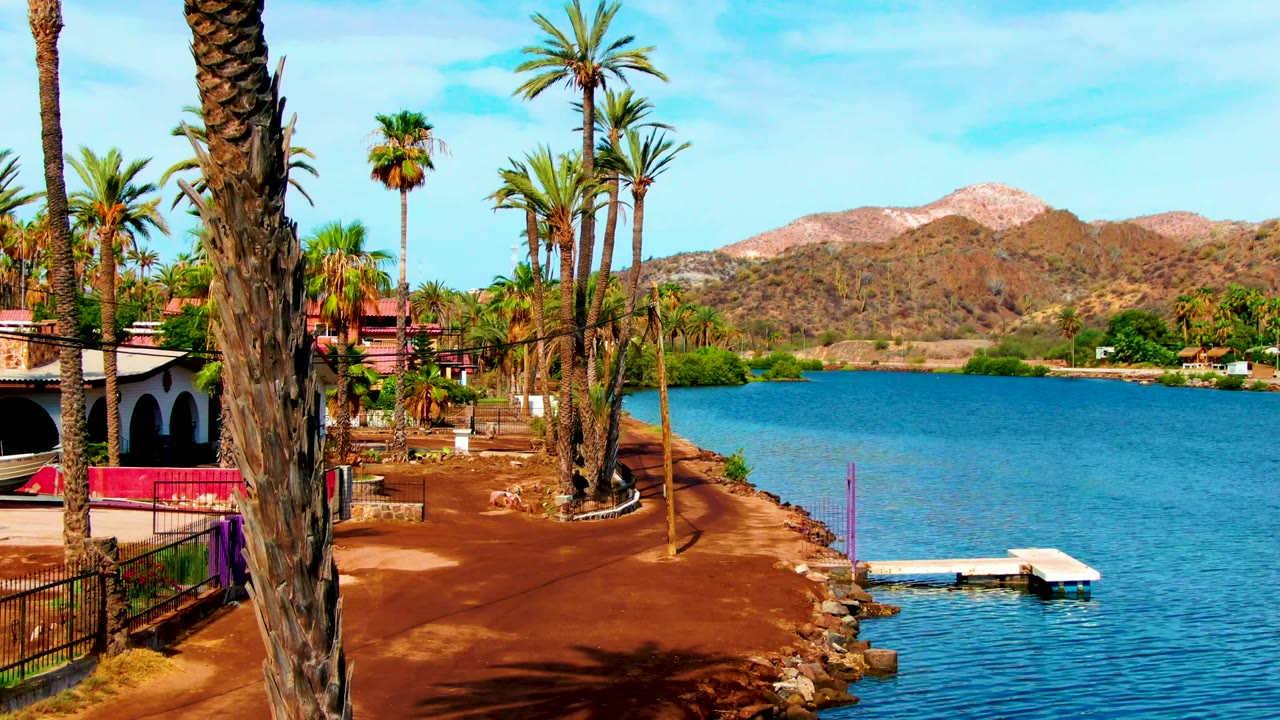 At 0:02 in Mulege, Baja California Sur, the composition opens slightly to the right, showing red shoreline, palms, calm water, a white dock, distant hills, and no visible people.