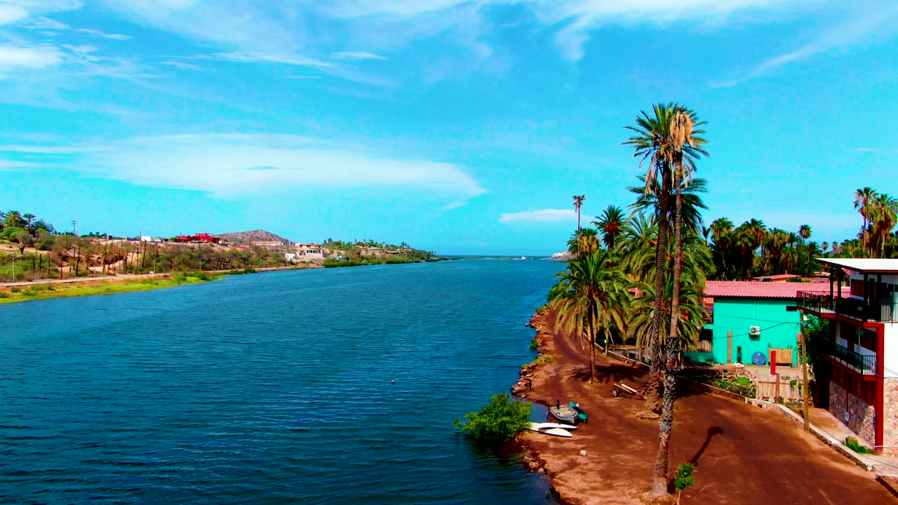 At 0:02, the Mulege river remains centered with fine ripples, a palm cluster on the right bank, turquoise and red building surfaces, low hills in the distance, and no visible people.