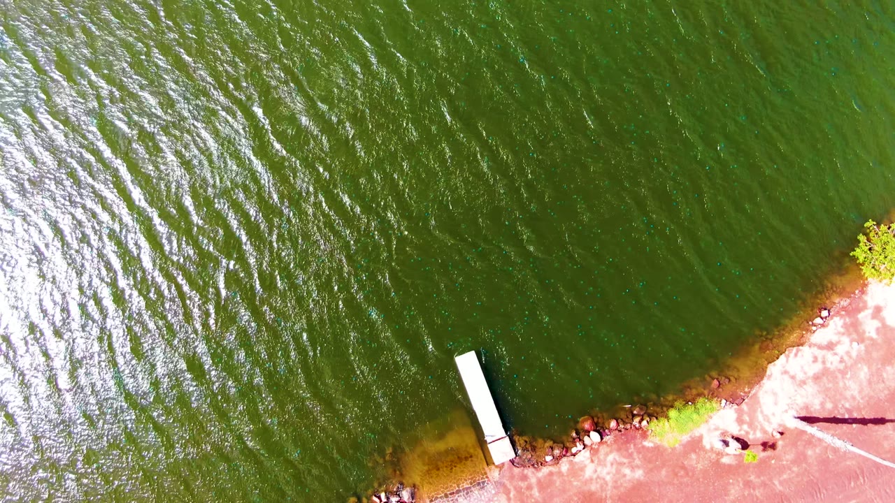 Overhead Mulege riverbank view showing dark green water, the small white dock, a rocky red-brown shoreline, a long thin shadow across the shore, and no visible people.