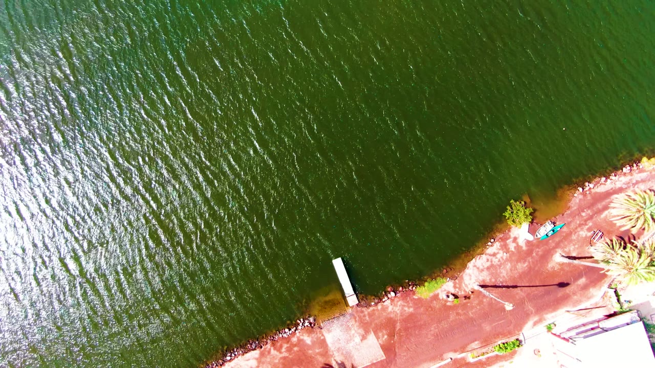 At 0:02, the Mulege shoreline sits lower in frame with green water dominating above, a white dock projecting from the bank, palms to the right, and distant small figures visible along the shore.