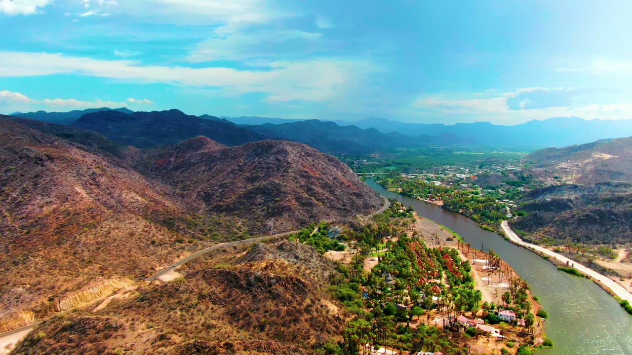 Early aerial frame over Mulege, Baja California Sur showing a greenish river curving past dry hills, roadside terrain, trees, scattered buildings, and no visible people.