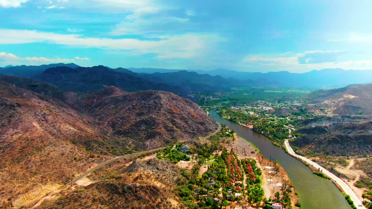 Aerial view in Mulege, Baja California Sur, Mexico, showing a green-brown river beside a dirt road, dry hills, clustered trees, small riverside structures, distant mountains, and no visible people.