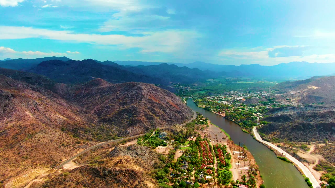 Wide aerial landscape with river, road, dry ridges, greenery, and distant mountains.