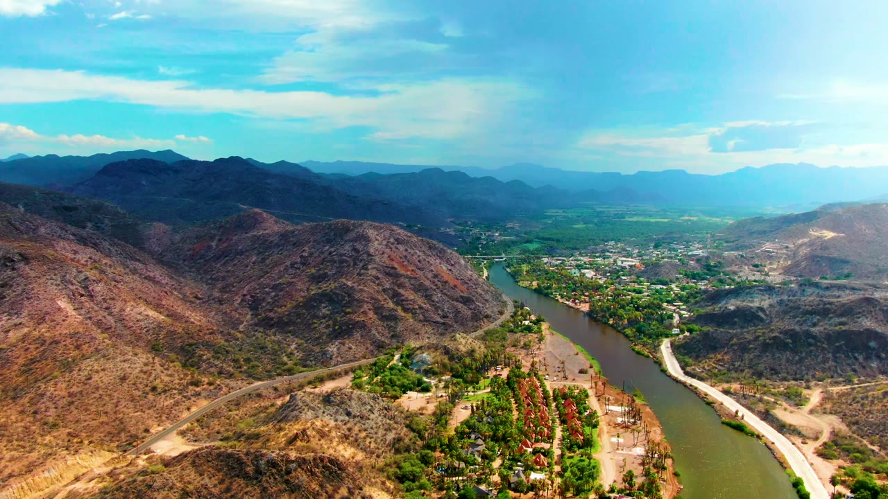 Drone-height view in Mulege showing a broad dry hillside in the foreground, a riverbank dirt road, green trees along the water, scattered buildings, hazy mountains on the horizon, and no visible people.