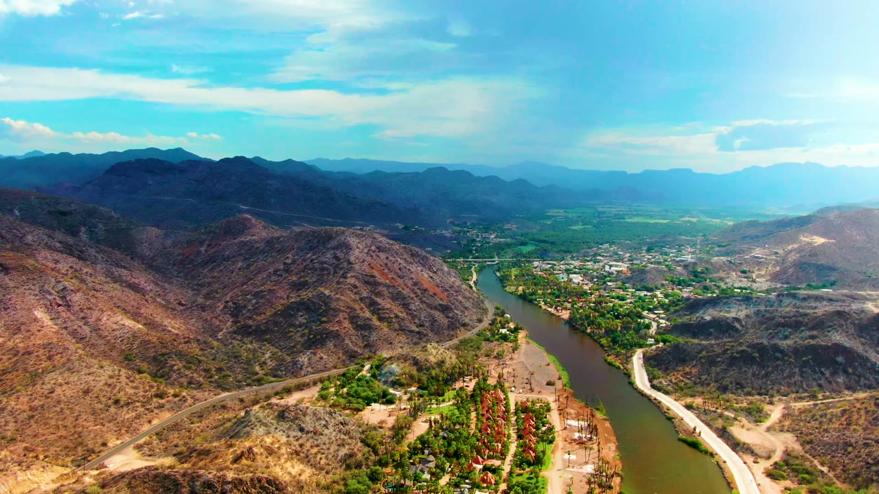 Wide aerial view over Mulege, Baja California Sur, with a river bending between dry rocky hills, a road along the bank, distant mountains, and no visible people.