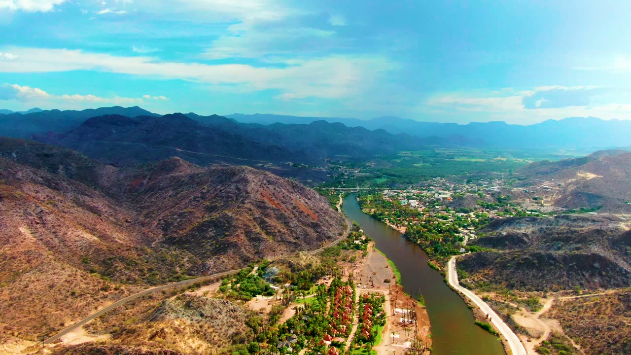A broad aerial view over Mulege with the river on the right side of the valley, reddish-brown slopes in front, green fields beyond, and no visible people.