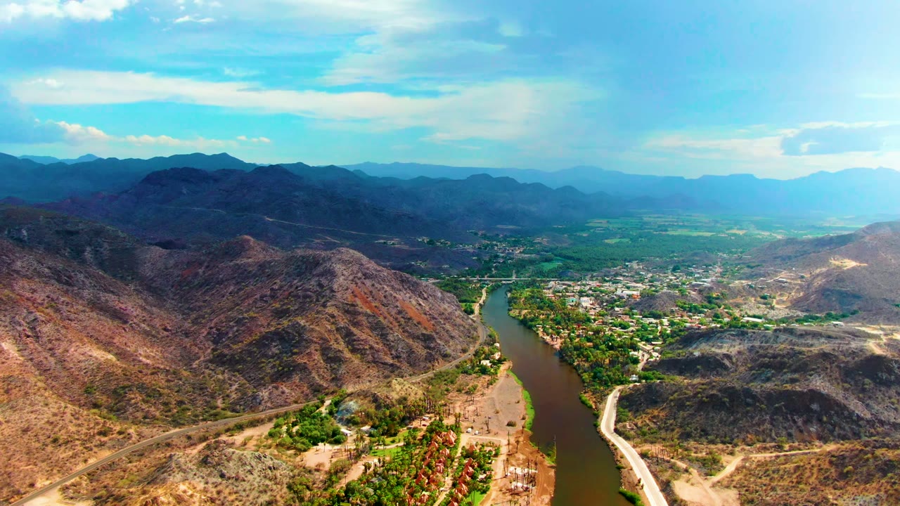 Aerial view over Mulege, Baja California Sur, with a winding brown-green river, dry hills, roads, distant mountains, blue sky, and no visible people.