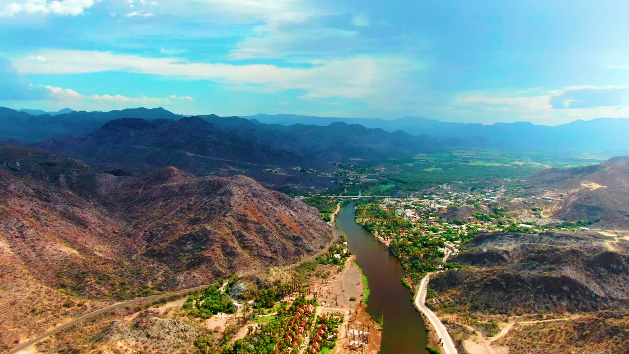 Aerial frame in Mulege, Baja California Sur, showing the river slightly right of center, green riverbank trees, rocky hills, distant ridges, and no visible people.