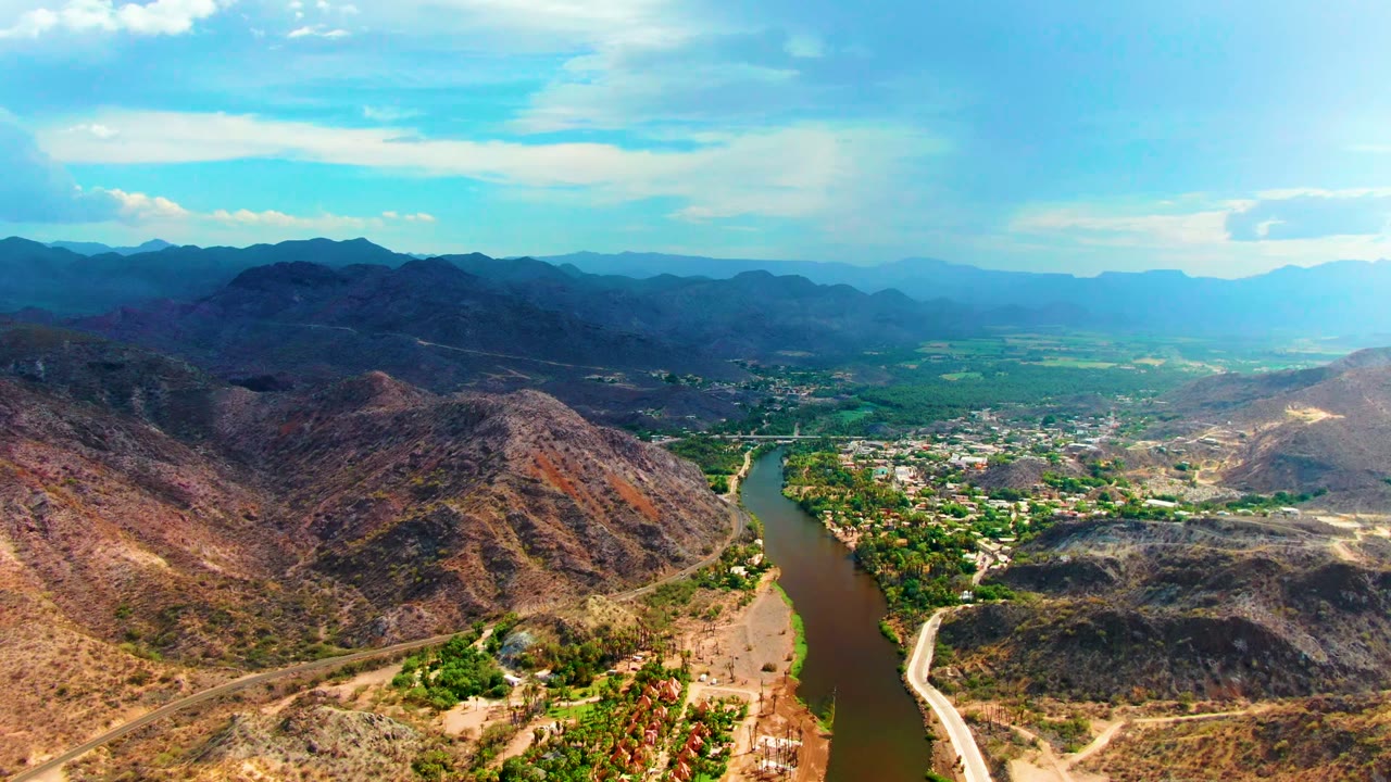 Wide aerial view of Mulege, Baja California Sur, with the river along the right side, clustered greenery and buildings below, dry mountain slopes, and no visible people.