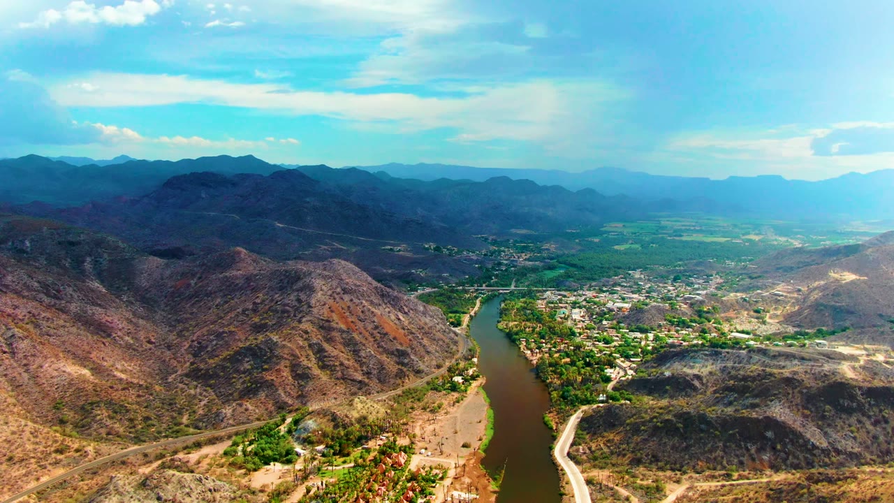 Opening aerial frame over Mulege, Baja California Sur, showing a brown winding river beside a small town, dry foreground hills, distant mountain ridges, bright clouds, and no visible people.