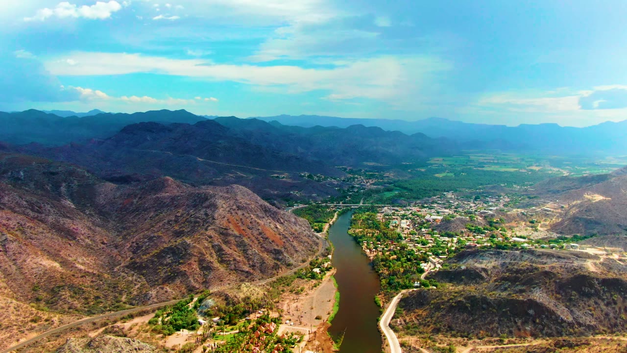 Wide Mulege aerial with a river bend at the lower left, a small settlement and green valley floor in the middle distance, rugged brown hills in front, hazy blue mountains behind, and no visible people.